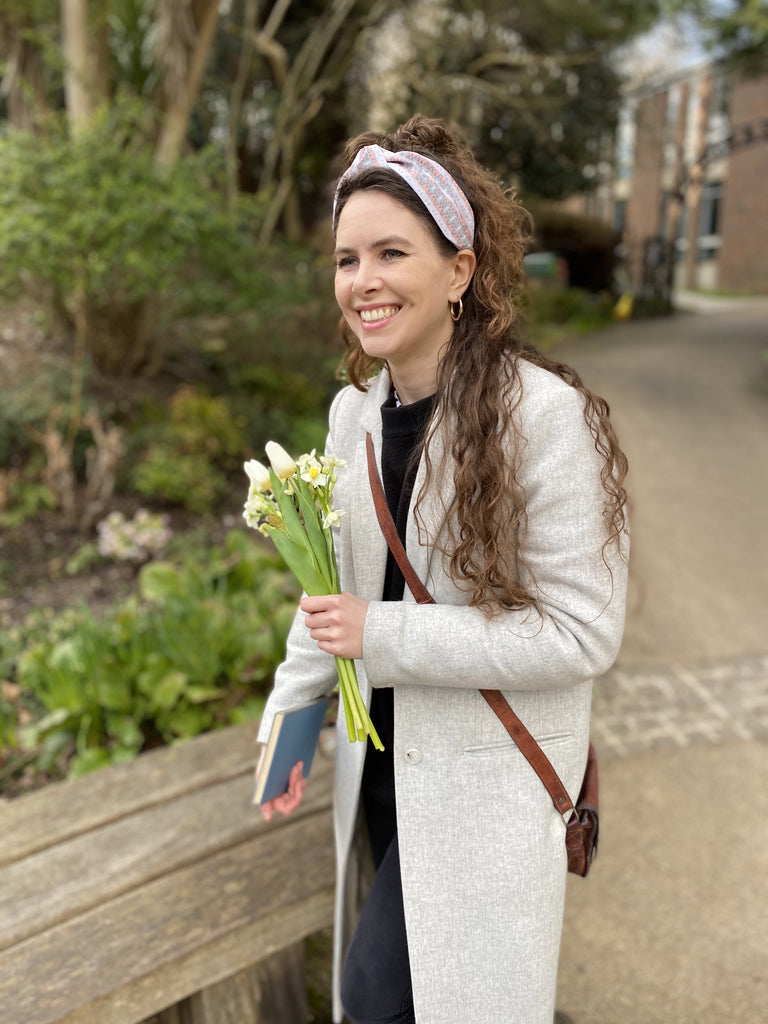 A brunette woman holding flowers and wearing a Kate Whyley wide, knotted headband in subtle pastel hues, called Cool Stride