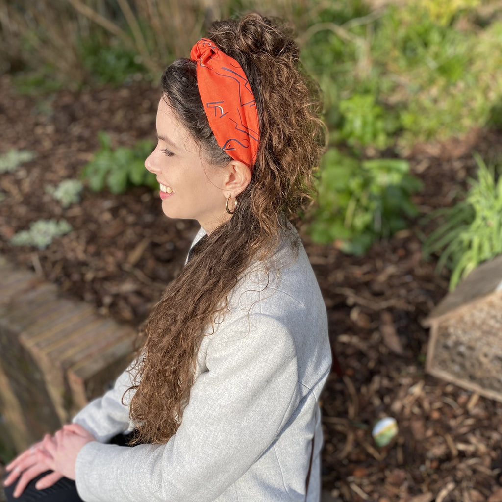 A brunette woman sitting on a wall, wearing a Kate Whyley wide, knotted headband in tangerine orange, with black and white caligraphy-like design