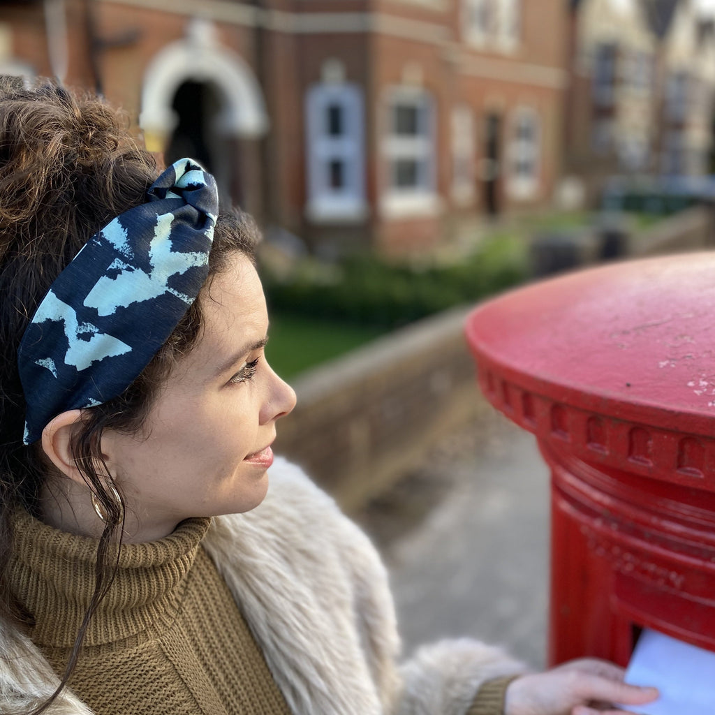 A brunette woman posting a letter, wearing a Kate Whyley wide, knotted headband with a mint green and navy pattern, called Studio