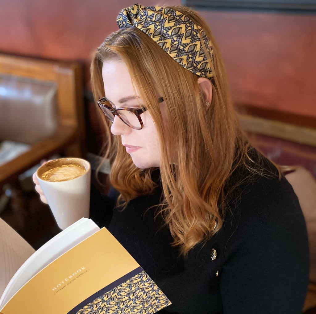 An auburn-haired woman holding a notebook and drinking a latte, wearing a Kate Whyley wide, knotted headband with a yellow, grey and black pattern, called Summer Leaf