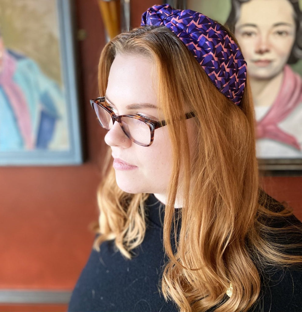 An auburn-haired woman wearing a Kate Whyley wide, knotted headband with a purple and pink pattern, called Nitesky